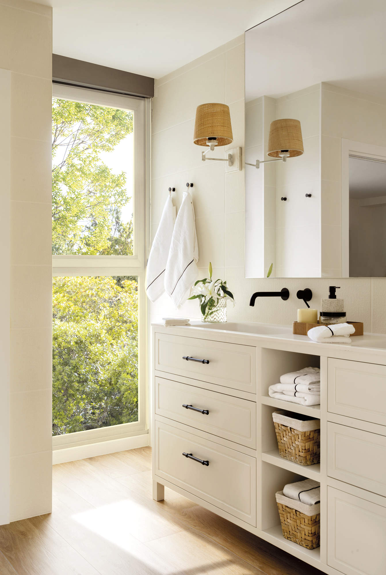 Baño con mueble de dos senos, grifería negra empotrada, espejo grande y aplique de pared, ventanal y suelo de madera
